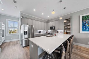 A modern kitchen with grey cabinetry, stainless steel appliances, a large island with bar stools, and a view of the outdoors through a window. - West Lagoon Cottages - Unit K | Beach Access | Private Balcony + Community Pool (Gulf Shores)