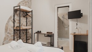 Une chambre d’hôtel avec un lit, une étagère en bois avec des serviettes, une petite table avec une tasse et une boîte, un téléviseur fixé au mur et un espace douche.