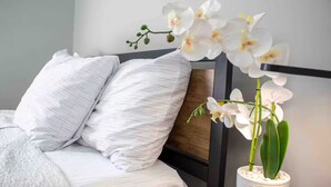 A bed with white pillows and a white bedspread, a wooden headboard, and a white vase with white orchids. - La Casita . (Winston-Salem)