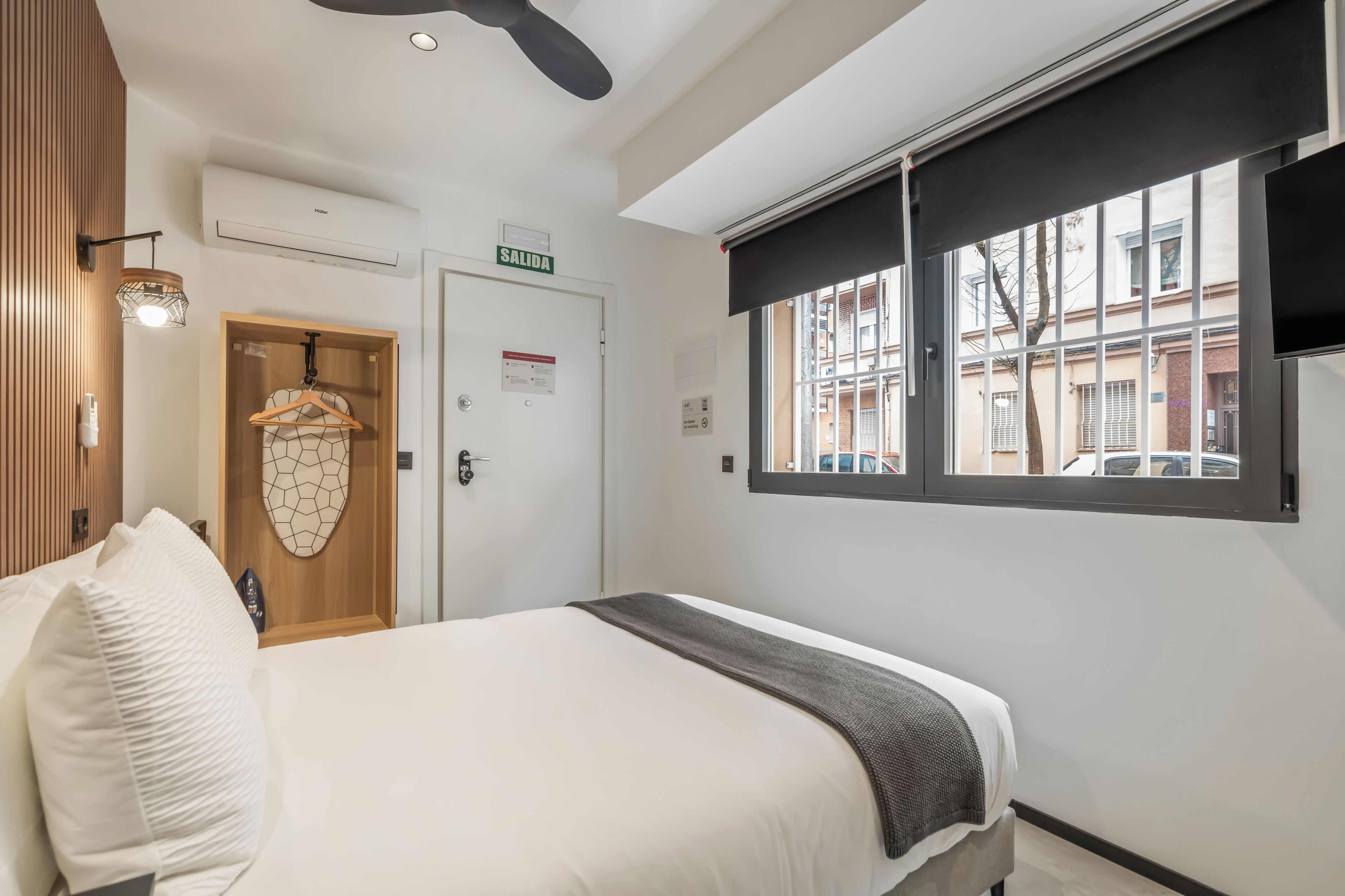 Ett hotellrum med en säng, en dörr med en skylt, en luftkonditionering, ett fönster med utsikt och en hängande lampa.