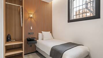 A small bedroom with a bed, a nightstand, a wall-mounted light, a window, and a wooden wardrobe.