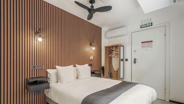 Une chambre d’hôtel équipée d’un lit, d’un ventilateur de plafond, d’une lampe fixée au mur, d’une table de chevet, d’une armoire et d’une porte portant l’inscription « SORTIE ».