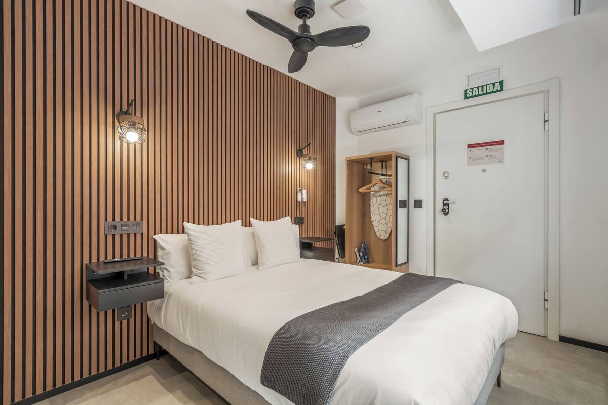 Habitació d'hotel amb un llit, un ventilador de sostre, una làmpada penjada a la paret, una taula de nit, un armari i una porta amb la inscripció 'SALIDA'.