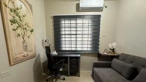 Une pièce comprenant un bureau, une chaise, un canapé, une fenêtre avec des stores, un climatiseur fixé au mur et une plante décorative.
