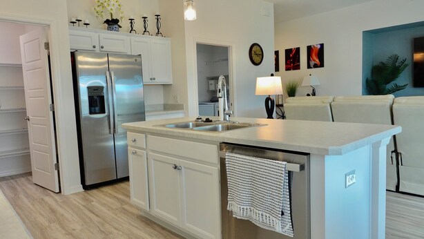 A modern kitchen with a stainless steel refrigerator, a built-in sink, and white cabinetry. There is a small island with a towel draped over it. - Beautiful 3/2 Designer Home with Golf Cart & Dog Friendly – Village of Dabney (The Villages)