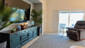 A modern living room with a flat-screen TV, a dark blue cabinet, a large potted plant, a brown armchair, and a glass coffee table. - Beautiful 3/2 Designer Home with Golf Cart & Dog Friendly – Village of Dabney (The Villages)