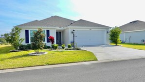 A two-story residential house with a white garage door, a front yard with a lawn and a small tree, and a driveway leading to the garage. - Beautiful 3/2 Designer Home with Golf Cart & Dog Friendly – Village of Dabney (The Villages)