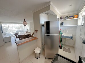 A modern kitchen with a stainless steel refrigerator, a dining area with wicker chairs, and a view through a large window. - Enseada Mutá (Porto Seguro)