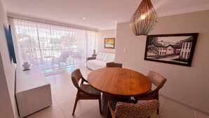 A dining area with a round wooden table, wicker chairs, a white sofa, and a large window with sheer curtains. - Enseada Mutá (Porto Seguro)