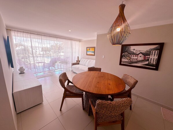 A dining area with a round wooden table, wicker chairs, a white sofa, and a large window with sheer curtains. - Enseada Mutá (Porto Seguro)