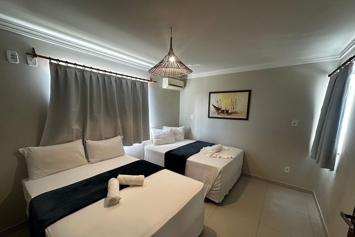 A hotel room with a bed, a smaller bed, a nightstand, a lamp, a painting, and a window with curtains.