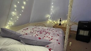 Ein Schlafzimmer mit einem mit Rosenblüten bedeckten Bett, einem Nachttisch mit Flasche und Kerzen sowie Lichterketten an der Wand.