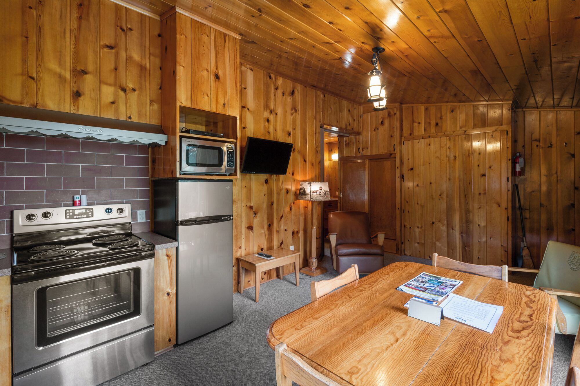 A compact kitchen with a stove, microwave, and refrigerator. There is a wooden dining table with chairs and a small table with a lamp.