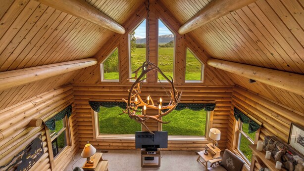 L’intérieur d’une cabane en rondins, avec un plafond haut, de grandes fenêtres, une cheminée et un lustre.