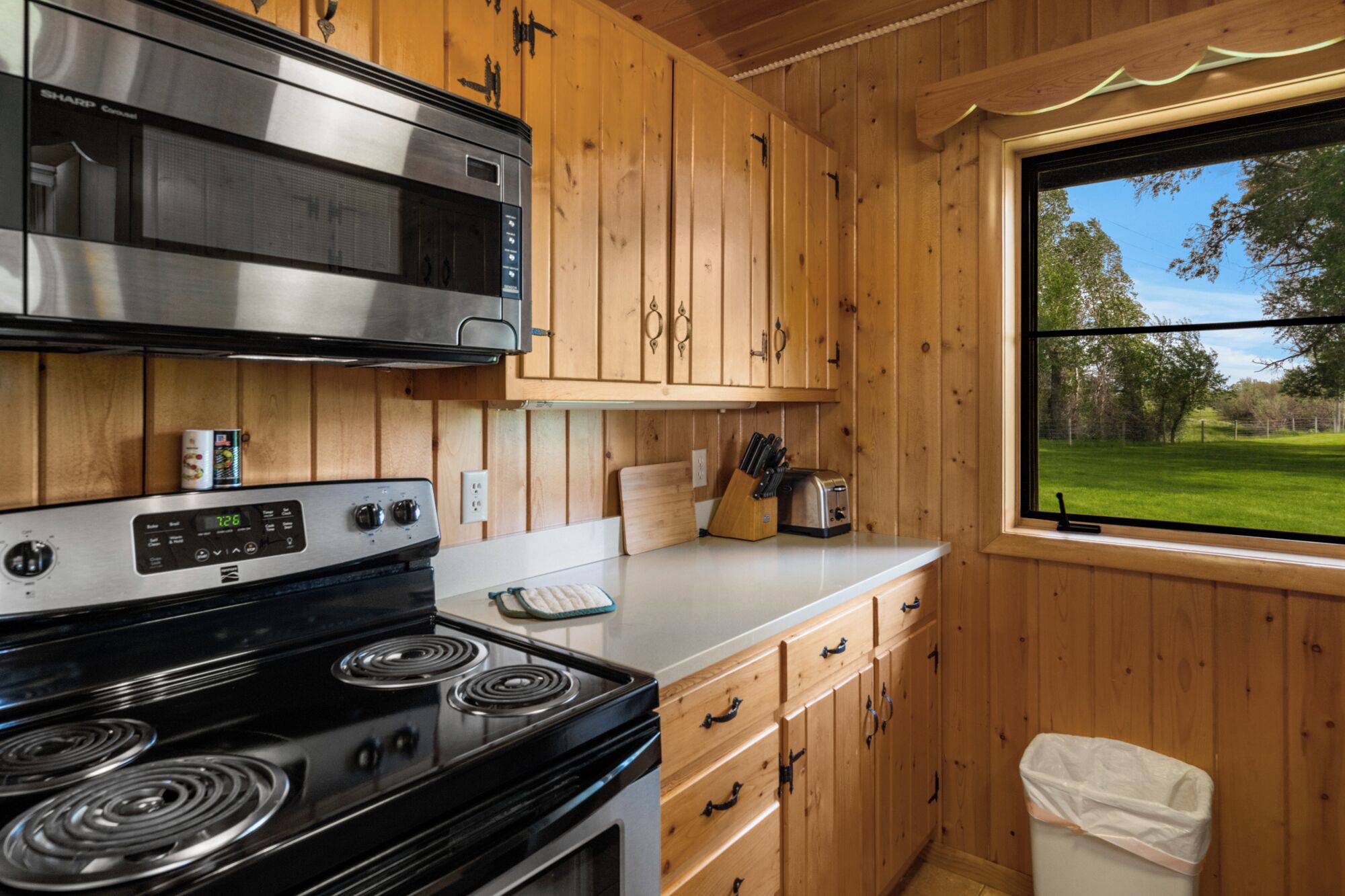 Une cuisine avec des meubles en bois, un four à micro-ondes en acier inoxydable, une cuisinière et une fenêtre donnant sur un champ verdoyant.