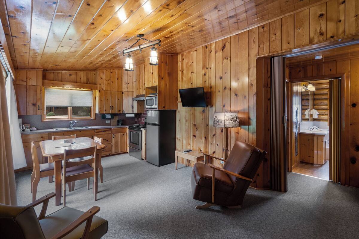 Ein gemütliches Blockhaus-Interieur mit Holzwänden und -böden, einem Essbereich mit Tisch und Stühlen, einer Küche mit Geräten und einer Sitzecke mit einem Sessel.