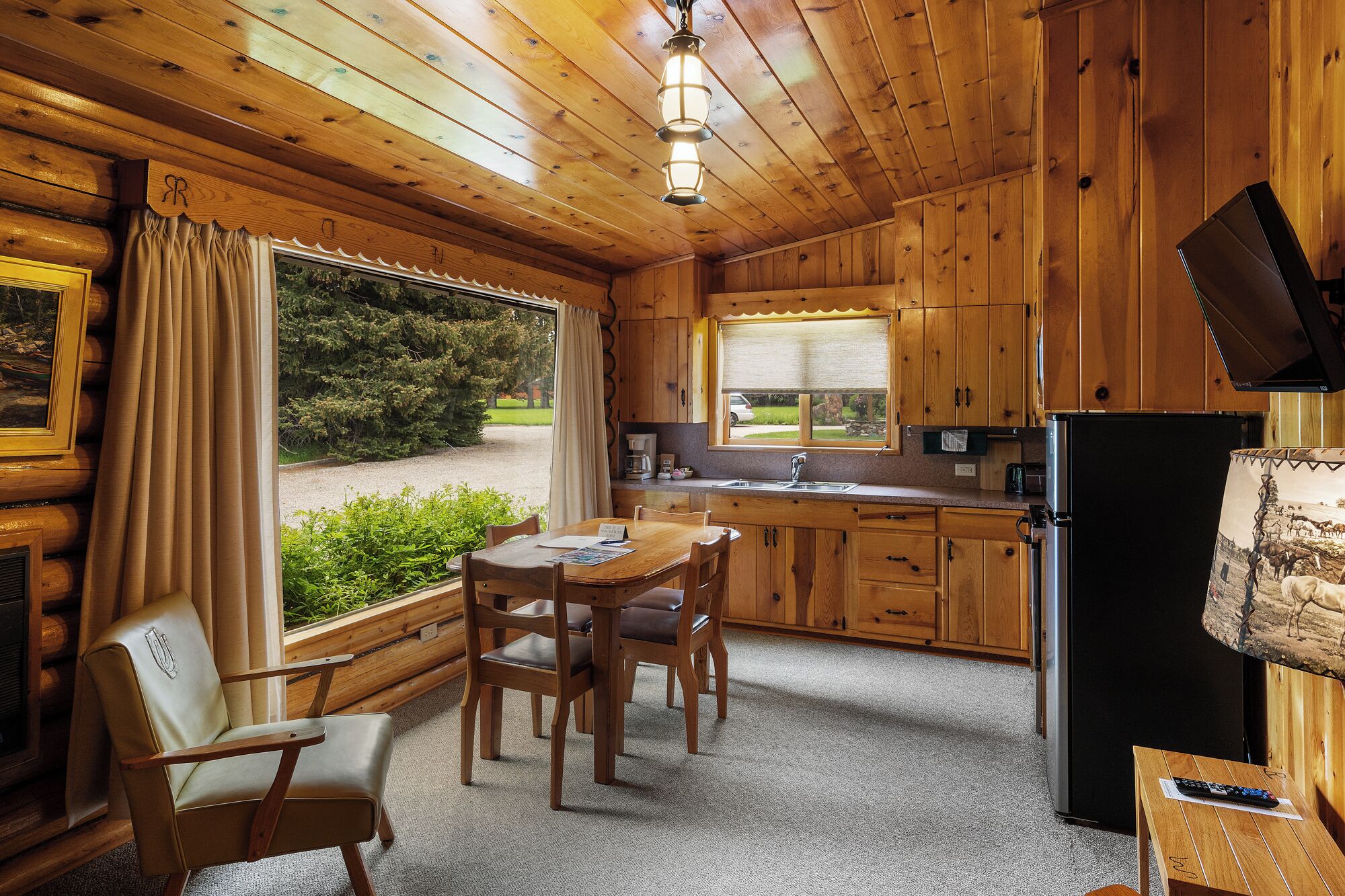 Una cocina acogedora estilo cabaña, con armarios de madera, un comedor con mesa y sillas, un refrigerador y una ventana con vistas a la vegetación.
