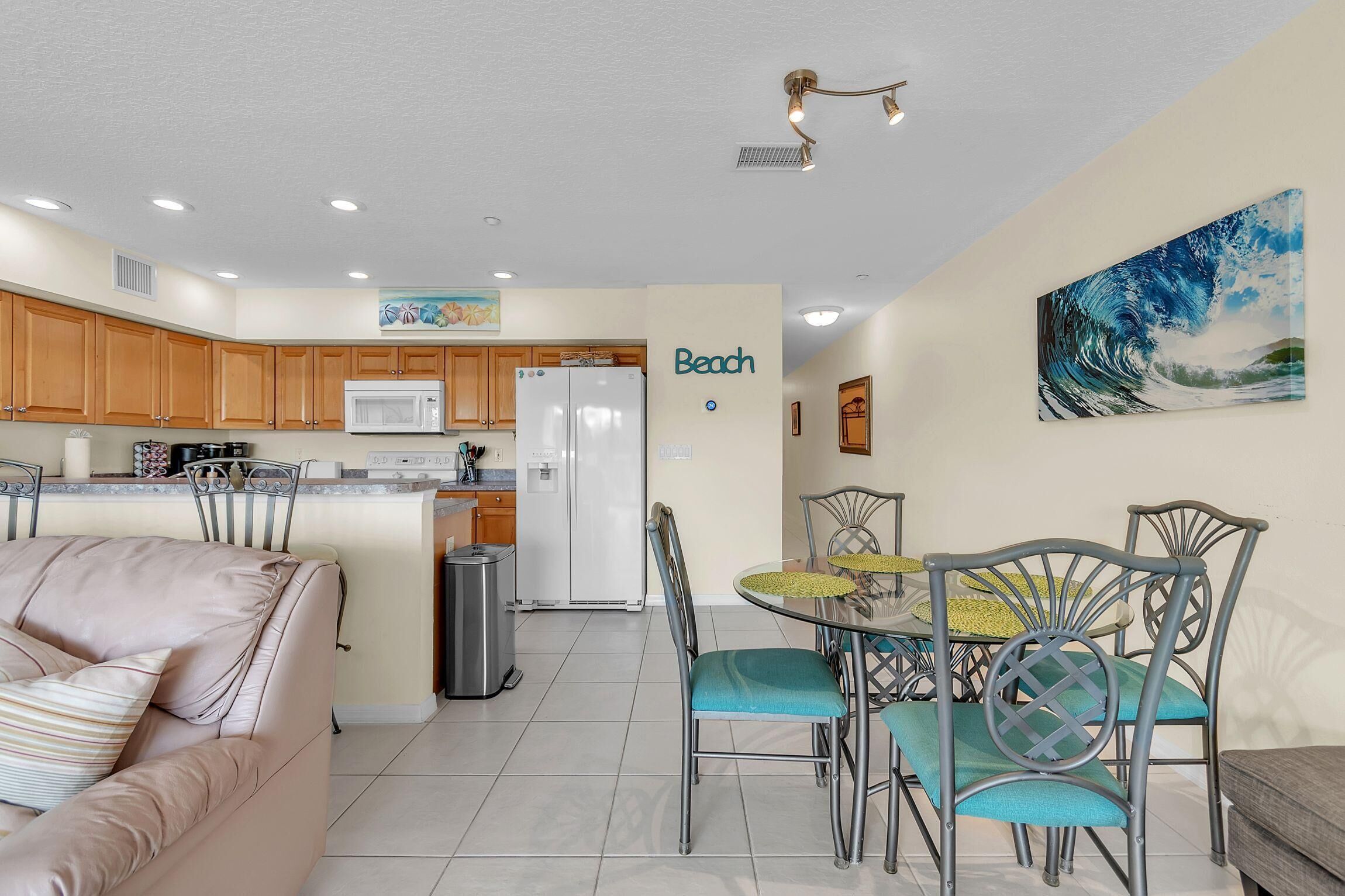 A modern kitchen with wooden cabinets, a white refrigerator, and a dining area with a glass table and metal chairs.