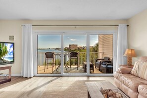 A living room with a large sliding glass door leading to an outdoor balcony with a view of a body of water and buildings. - Captain's Cove #203 (Indian Shores)