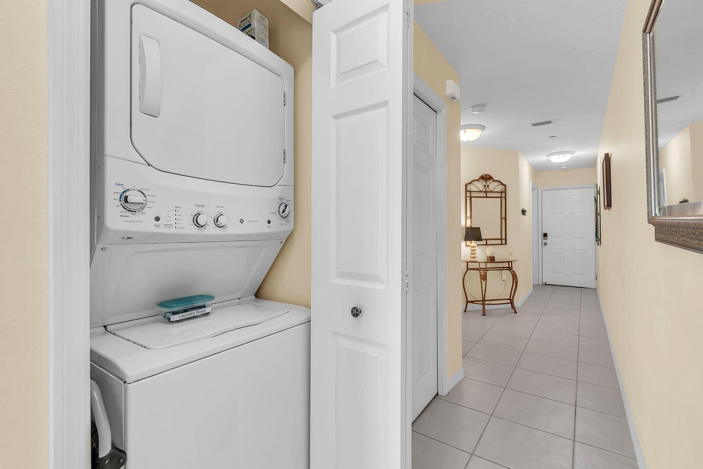 A laundry room with a front-loading washer and dryer, a small table with a mirror, and a door leading to another room.