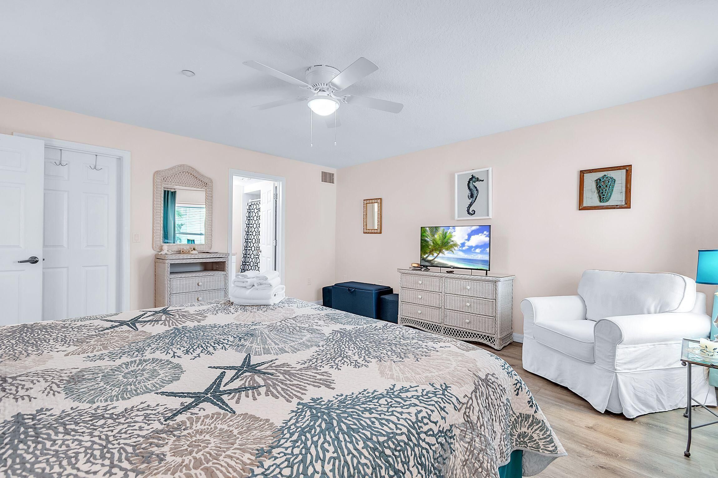 A bedroom with a bed, a ceiling fan, a dresser, a white armchair, and a television.