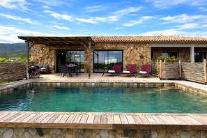 Un espace aménagé au bord de la piscine, avec un mur en pierre, une terrasse couverte et une vue sur les montagnes.
