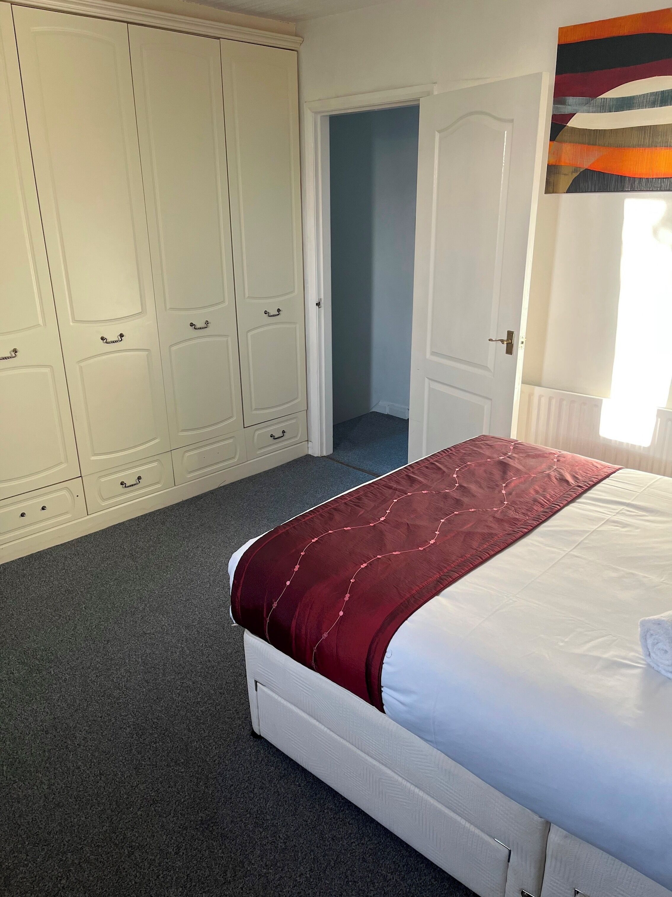 A bedroom with a large white wardrobe, a bed with a red blanket, and a view of a hallway.