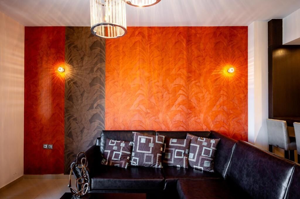 A modern living room with a black leather sofa, patterned cushions, and a textured orange wall.