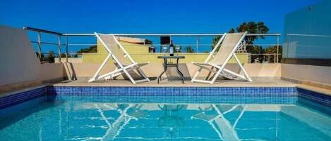 Ein klares, blaues Schwimmbecken mit zwei Liegestühlen und einem Tisch, umgeben von einem Glasgeländer.