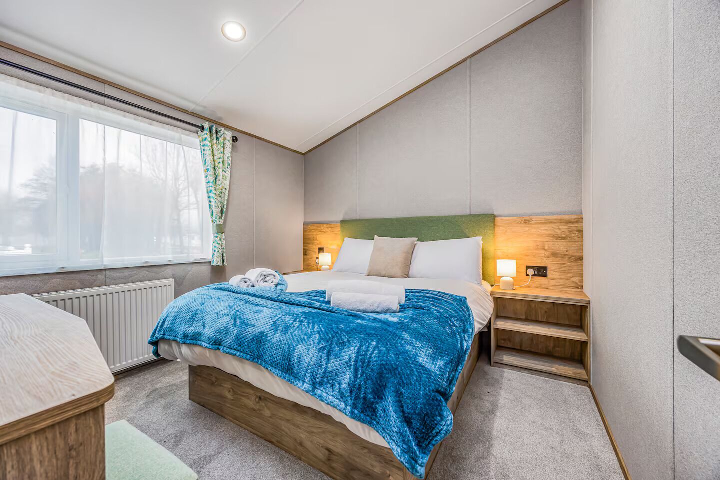 Een moderne hotelkamer met een groot bed, een houten hoofdeinde, een nachtkastje met een lamp, en uitzicht op de omgeving door een raam.