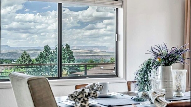 Uma área de refeições com uma mesa posta para dois, uma janela com vista para uma paisagem montanhosa e um vaso com flores na mesa.