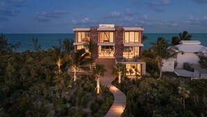 A modern two-story house with a wooden deck leading to the entrance, surrounded by tropical vegetation. - Beachfront Villa w/ Pool & Ocean Views (Long Bay)