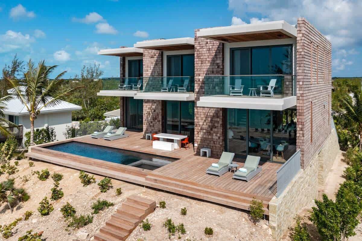 A modern two-story house with a swimming pool, outdoor seating, and a view of the ocean.