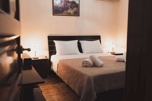 Een net opgemaakt bed met een bruine deken, twee witte kussens en een opgerolde handdoek aan de zijkant. Er staat een nachtkastje met een lamp bij, en een klein tafeltje met een boek erop. Een schilderij hangt aan de muur boven het bed.