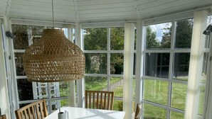 A conservatory with a white table and chairs, a woven lamp, and large windows offering a view of a garden. - 4 Star Holiday Home in Hals (Hals)