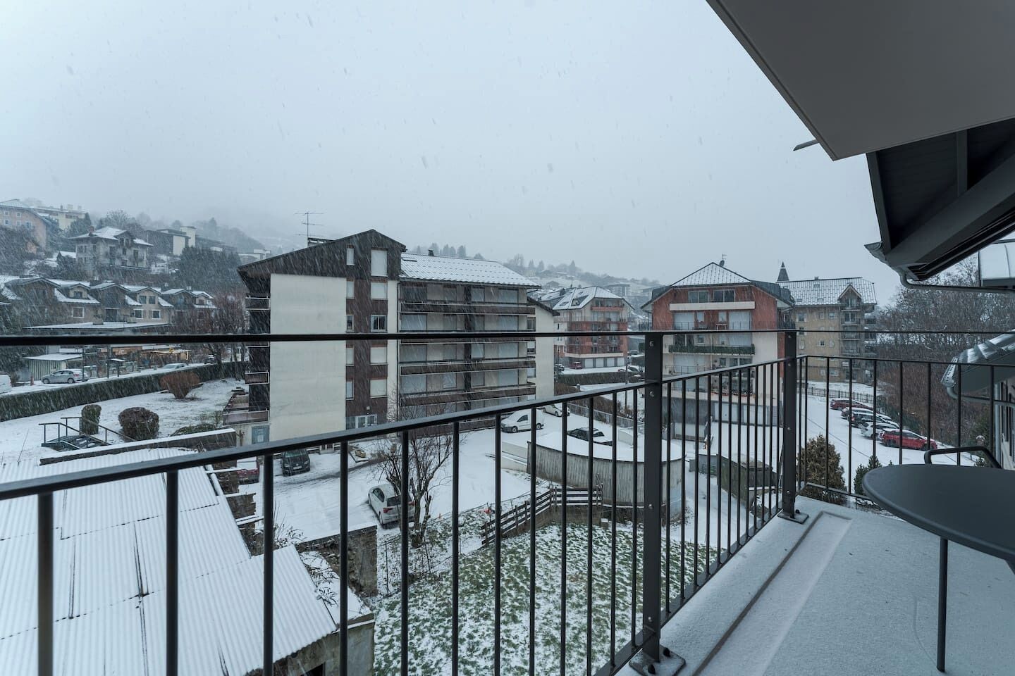Un barrio residencial nevado con edificios de varios pisos, un balcón con barandilla de metal y vistas a coches aparcados y una colina distante.