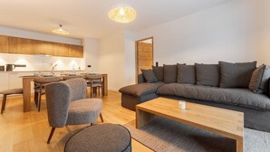 Een moderne woonkamer met een grijze bank, een houten salontafel en grijze fauteuils. In de achtergrond is een eethoek met een houten tafel en stoelen zichtbaar.