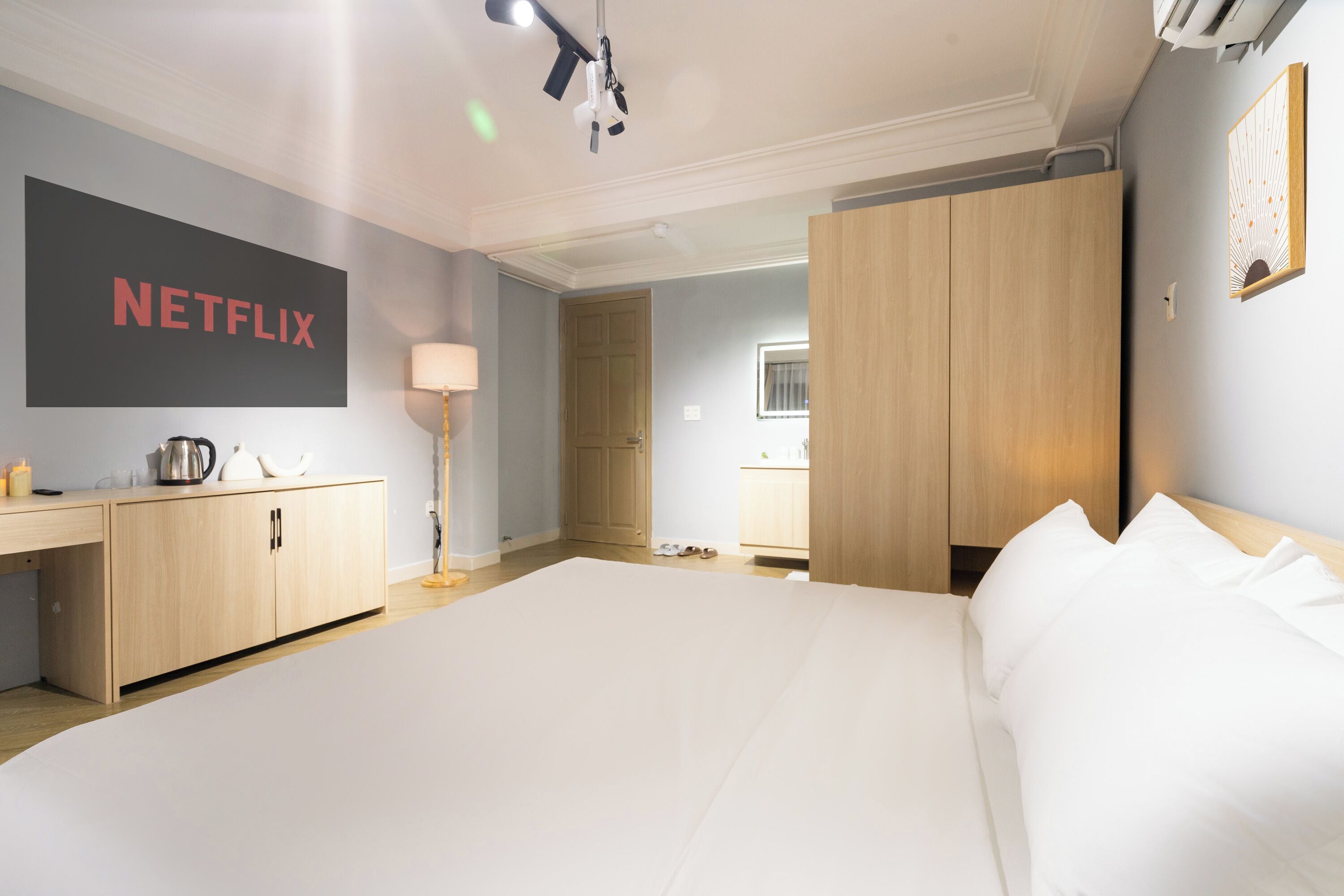 A modern hotel room with a large bed, a wooden wardrobe, a desk with a kettle, and a wall-mounted Netflix sign.
