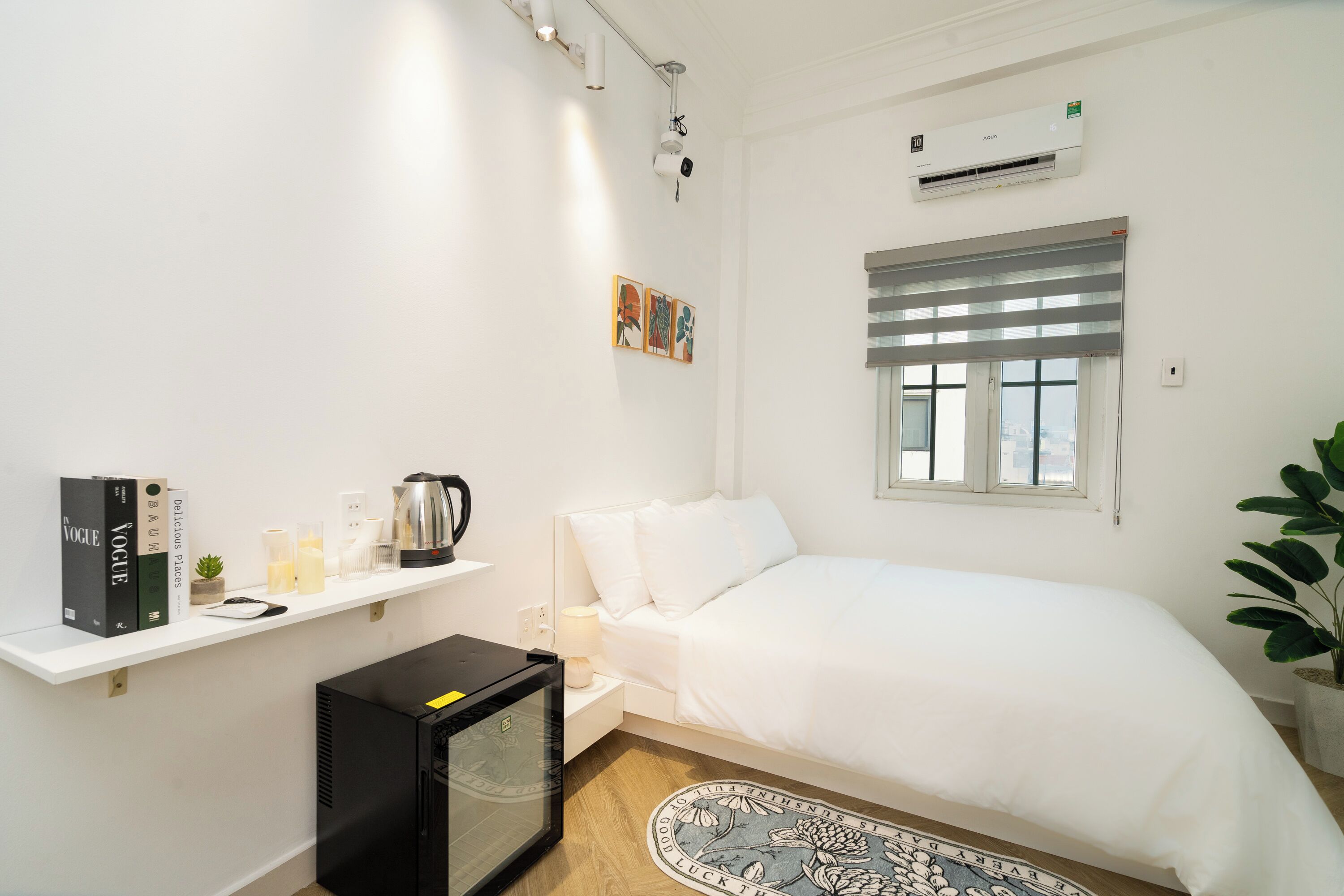 A small, clean room with a bed, a shelf with books and a coffee maker, a small table with a lamp, and a potted plant.