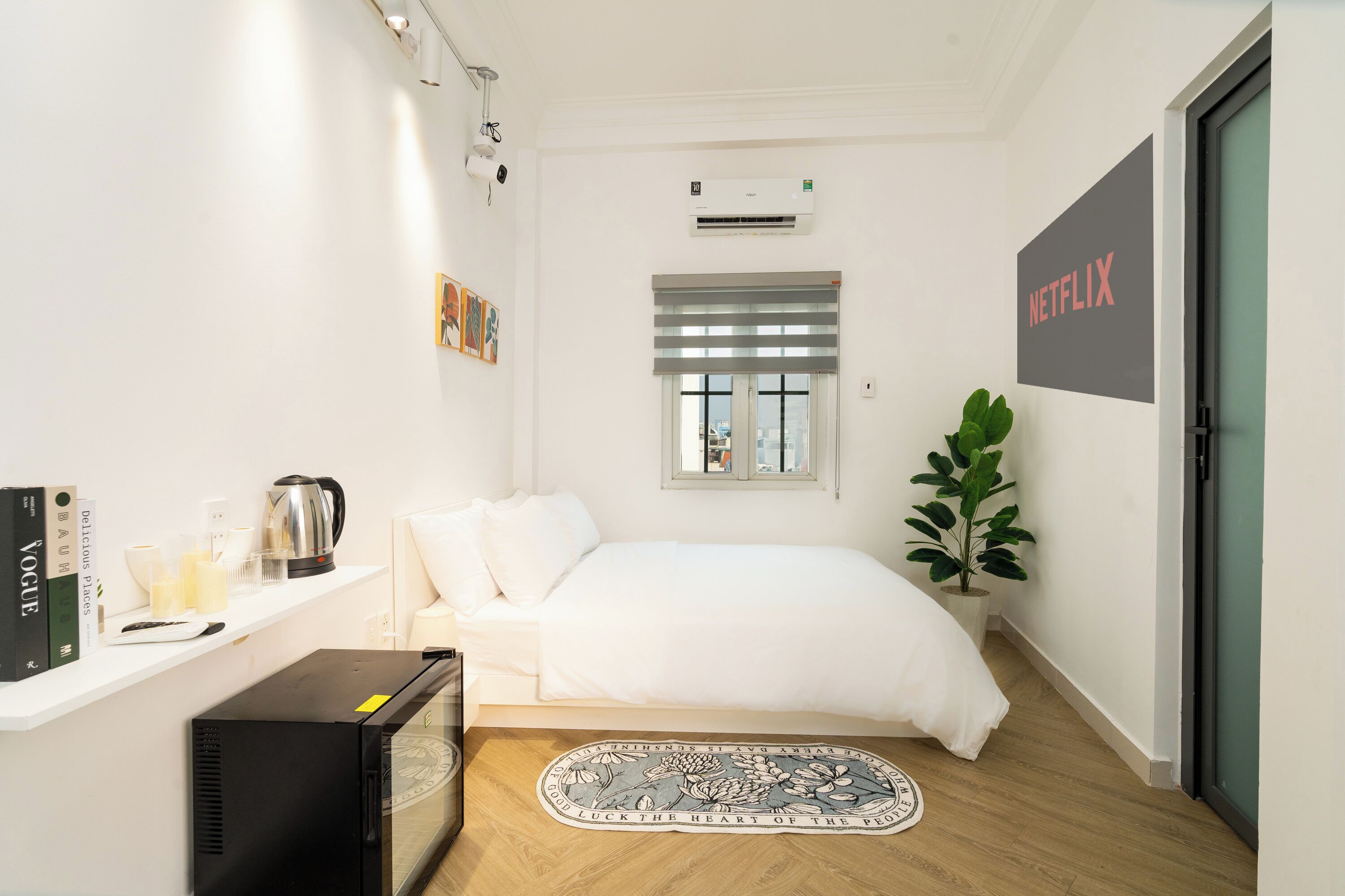 A modern bedroom with a bed, a small desk with a computer, a coffee maker, and a potted plant.