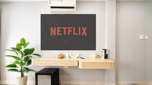 A modern room with a wooden floor, a potted plant, a small black refrigerator, a wooden desk with a mounted TV displaying 'NETFLIX', and a pair of shoes by the wall. - Chill Hill Hotel & Home (Ho Chi Minh City)