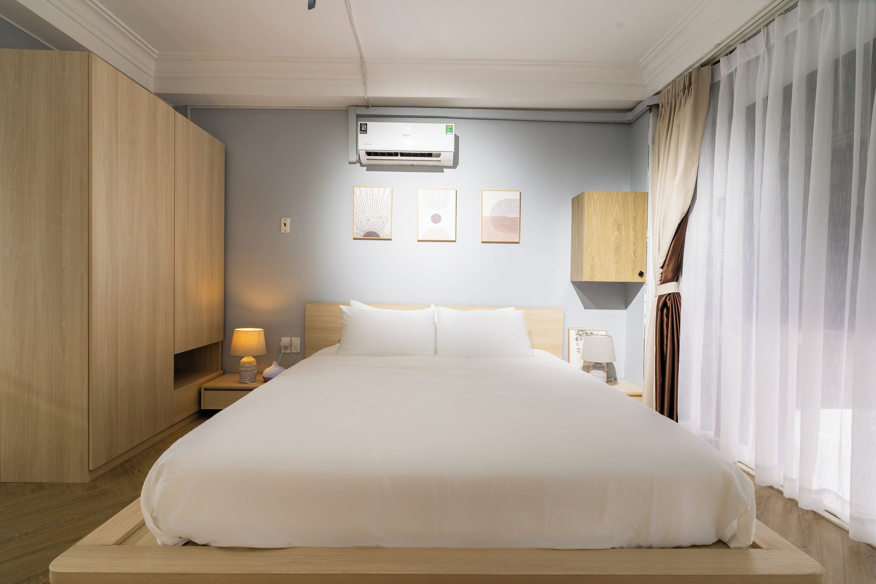 A hotel room with a large bed, a nightstand with a lamp, an air conditioning unit, and a window with curtains.