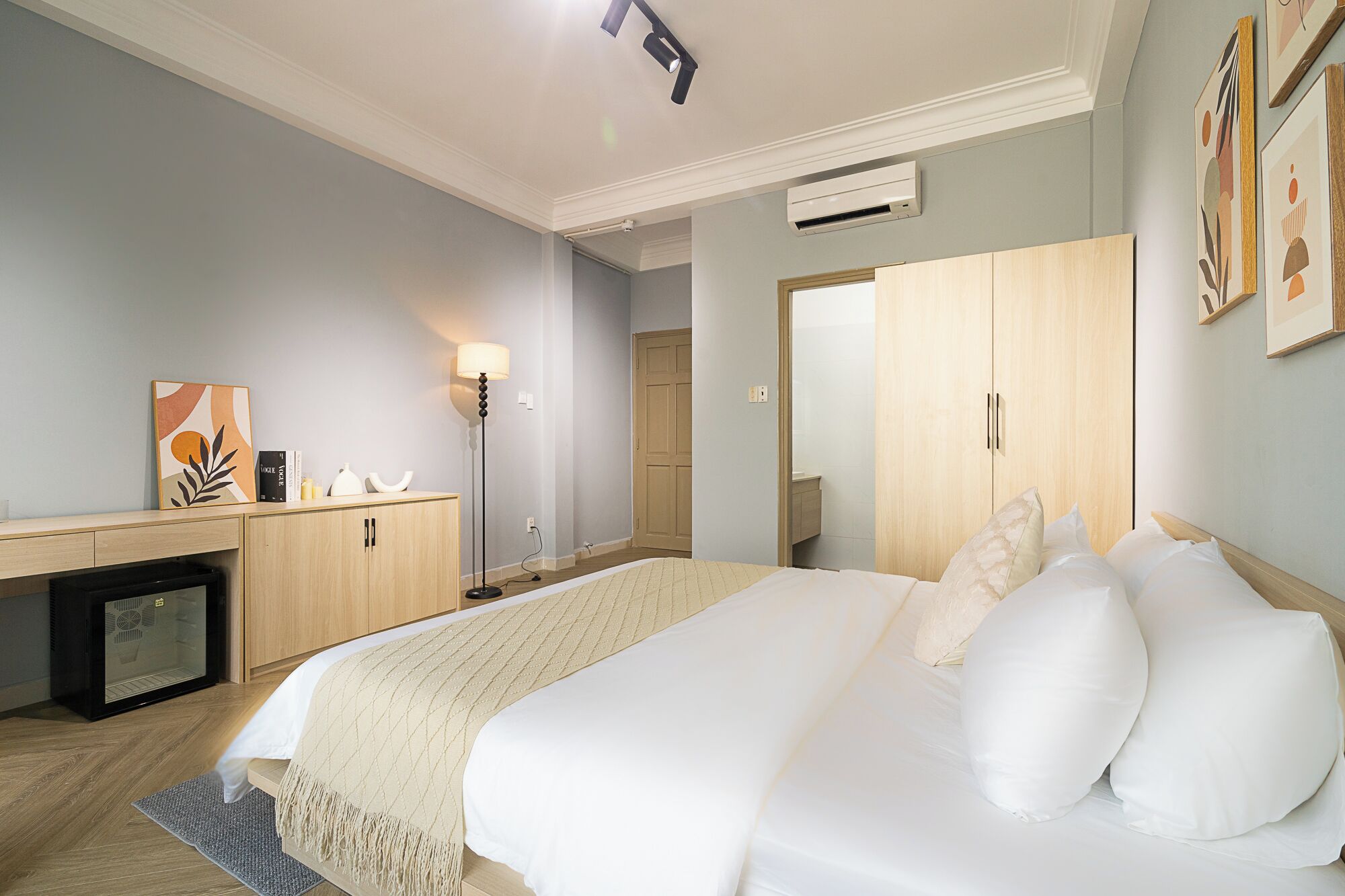A hotel room with a bed, bedside table, lamp, wardrobe, and artwork on the wall.