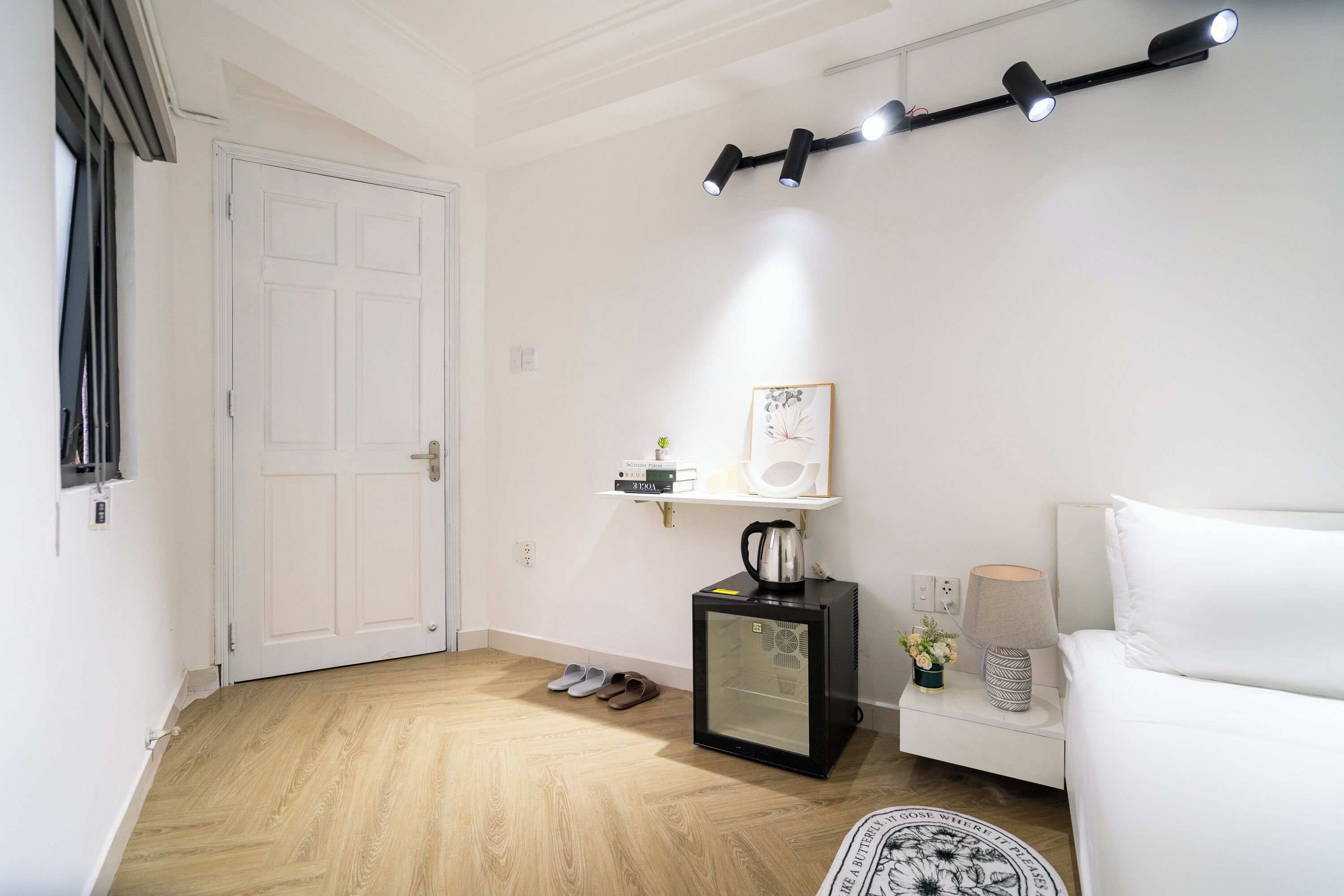 Une pièce avec une porte blanche, un petit réfrigérateur noir, un canapé blanc, une machine à café et un luminaire fixé au mur.