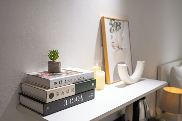 A stack of books on a white shelf, a small potted plant, a candle, and a framed artwork.