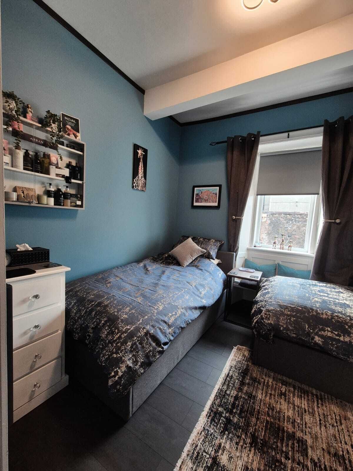 A bedroom with a bed, a nightstand, a dresser, a window with curtains, and a wall with shelves containing toiletries.