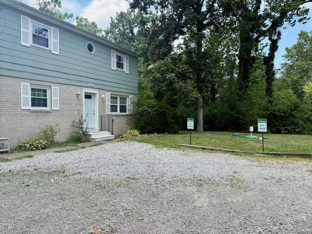A two-story building with a gravel driveway and a small front porch. There are two signs indicating guest parking.