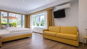 Une chambre d’hôtel avec un lit, un canapé jaune, un téléviseur fixé au mur et de grandes fenêtres avec des rideaux.