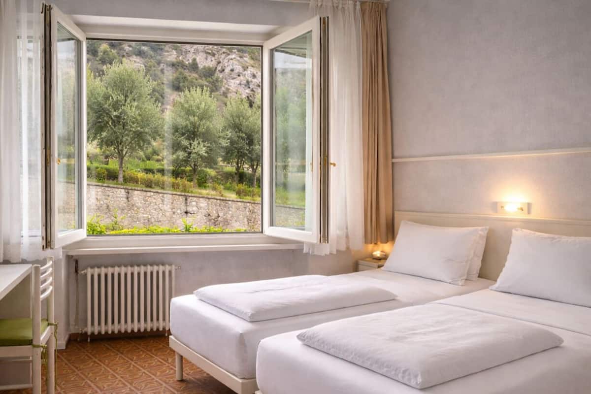 Un dormitorio con dos camas, una ventana que da a árboles y un muro de piedra, y un radiador.