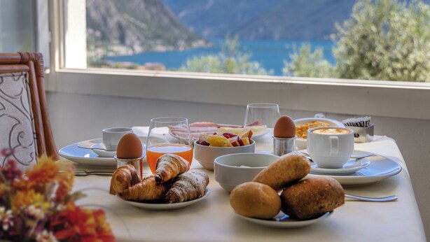 Une table de petit-déjeuner avec des croissants, des fruits, du café et une vue sur les montagnes et l’eau.